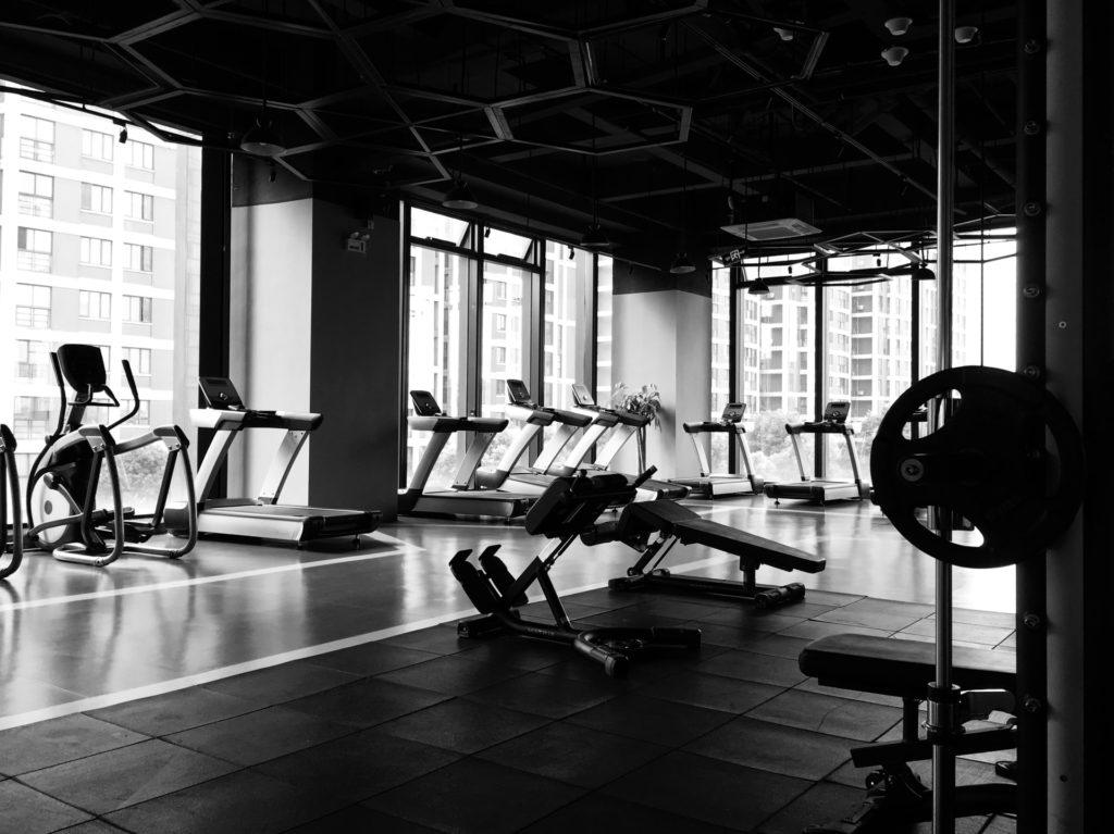 Espace cardio d'une salle de sport dans le fond (tapis de course, vélos) mais aussi un banc de musculation au mileu