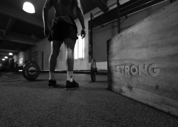 Personne devant une barre de musculation dans une salle de sport, photo en noir et blanc.