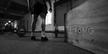 Personne devant une barre de musculation dans une salle de sport, photo en noir et blanc.