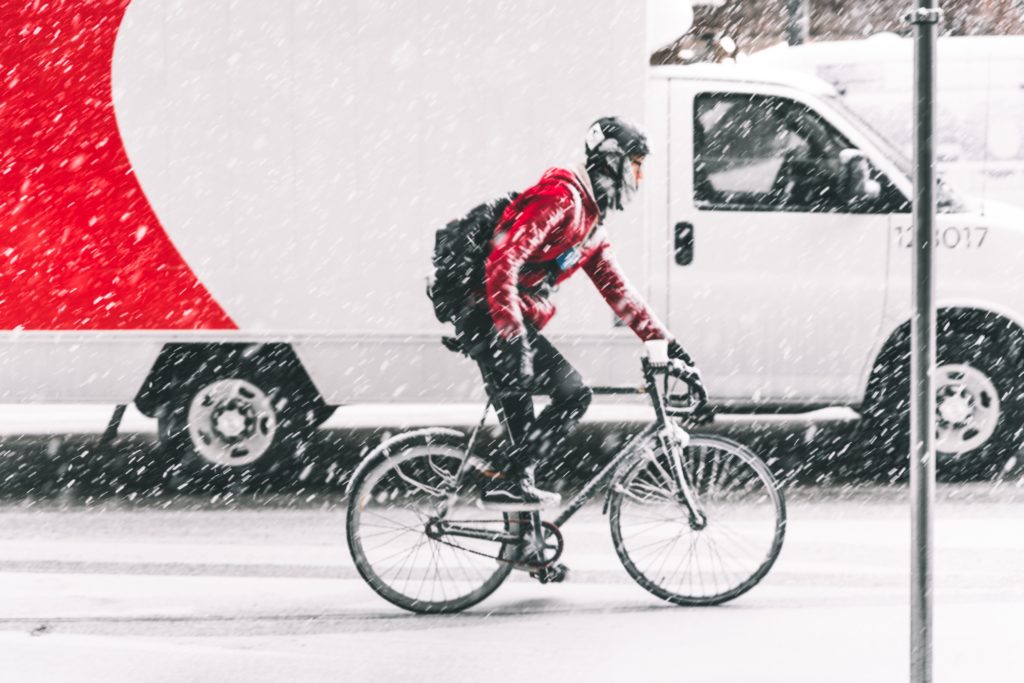 Cycliste avec un haut rouge circulant sur une route qui brave une tempête de neige. On aperçoit un camion en arrière-plan qui roule à côté du cycliste.