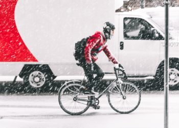 Cycliste avec un haut rouge circulant sur une route qui brave une tempête de neige. On aperçoit un camion en arrière-plan qui roule à côté du cycliste.