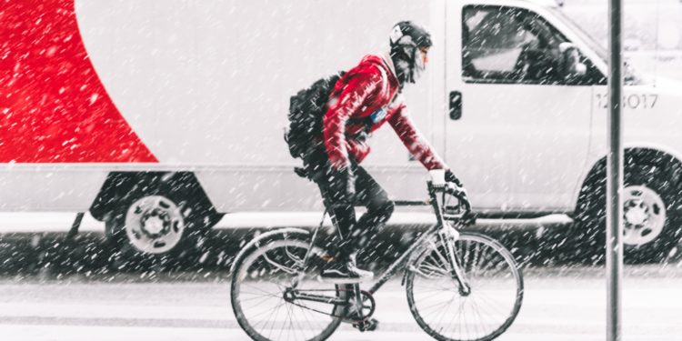 Cycliste avec un haut rouge circulant sur une route qui brave une tempête de neige. On aperçoit un camion en arrière-plan qui roule à côté du cycliste.