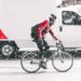 Cycliste avec un haut rouge circulant sur une route qui brave une tempête de neige. On aperçoit un camion en arrière-plan qui roule à côté du cycliste.