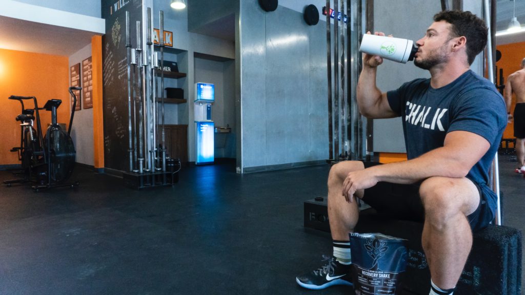 Un homme assit dans une salle de sport en train de boire un shaker protéiné.