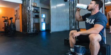 Un homme assit dans une salle de sport en train de boire un shaker protéiné.