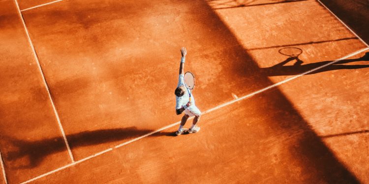 Court de tennis sur terre battue, un joueur est en train de faire un service, la balle est en l'air