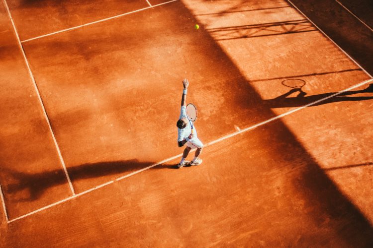 Court de tennis sur terre battue, un joueur est en train de faire un service, la balle est en l'air